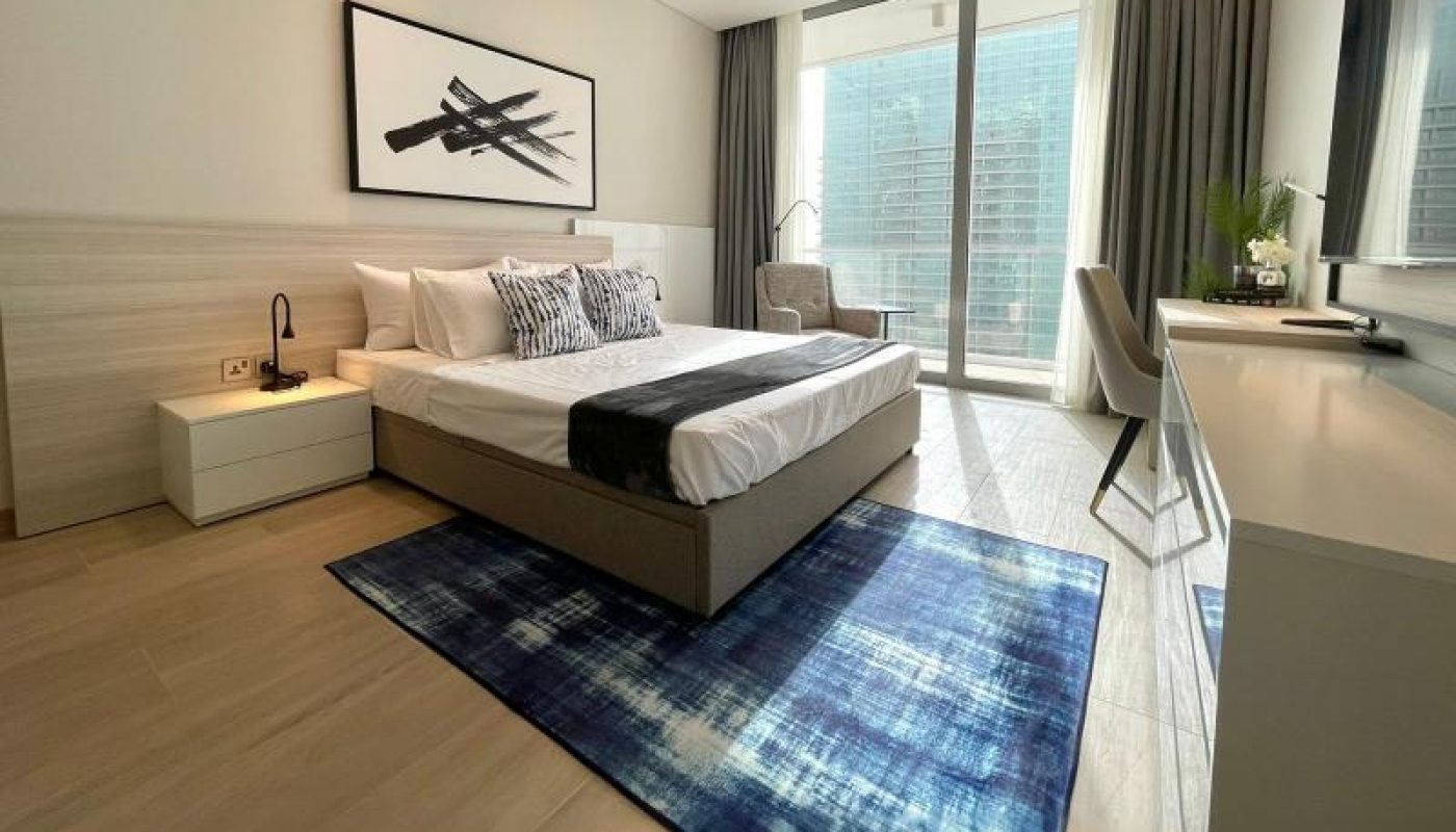 Modern bedroom with a double bed, white and blue decor, large window with city view, desk, chair, and wall art above the bed. Natural light brightens the room.