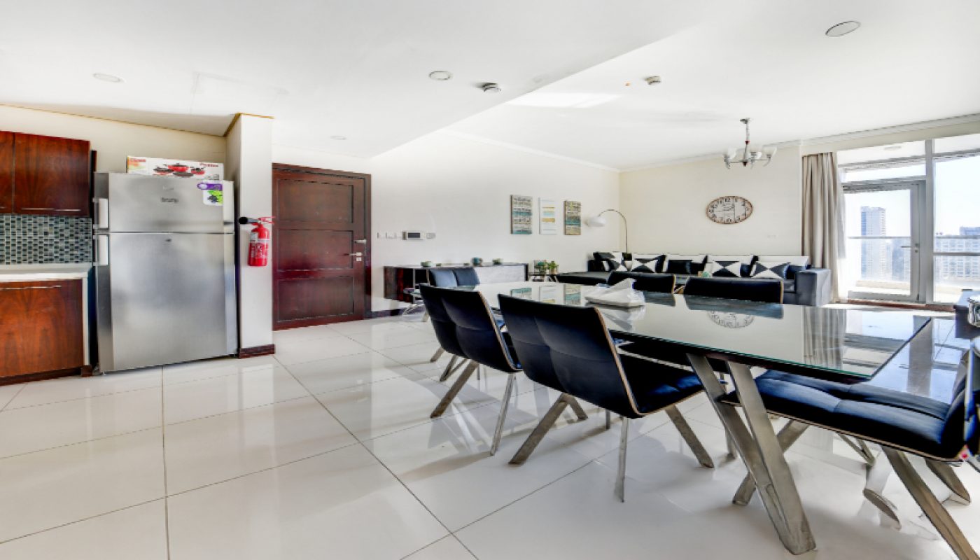 Modern open-plan kitchen, dining, and living area with tile flooring, glass dining table, black chairs, large windows, and stainless steel refrigerator.
