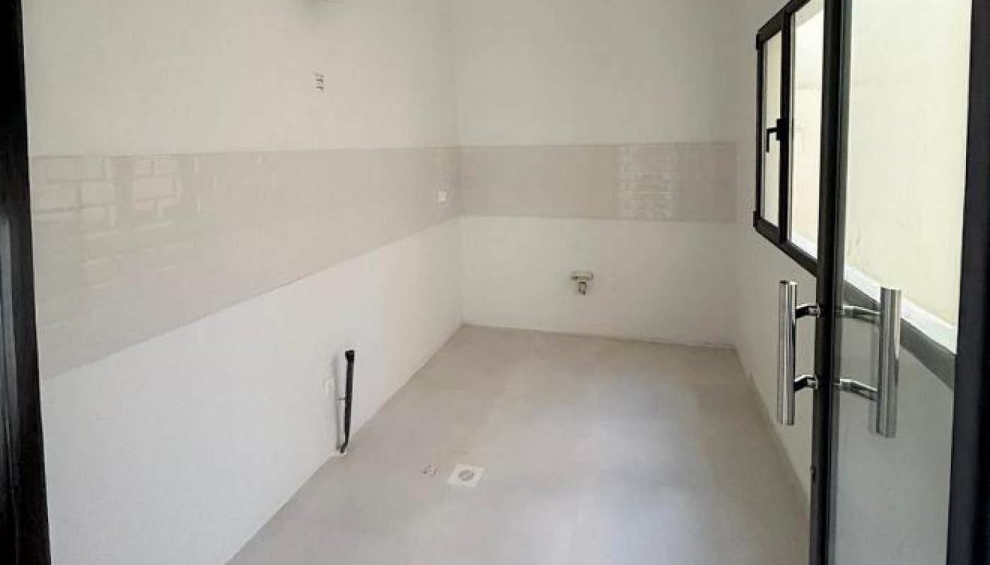 Empty, unfurnished room with white walls, tiled floor, a window, and a glass door. Exposed pipes and vents suggest it is intended to be a kitchen or utility area.
