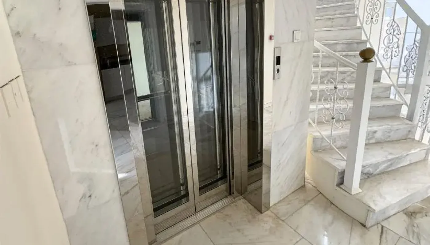 A modern elevator with mirrored doors is positioned next to a white marble staircase in a well-lit interior with marble walls and flooring.