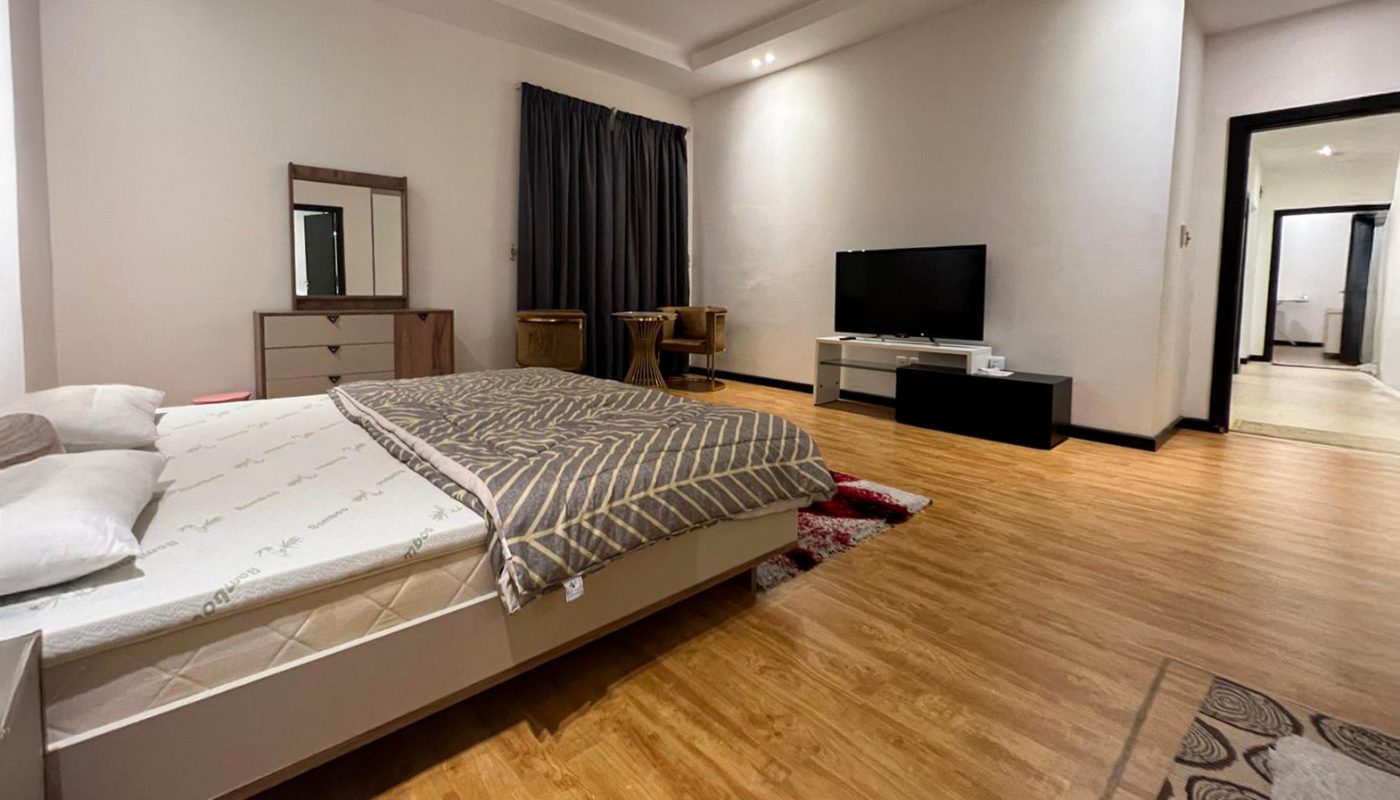 Spacious bedroom with wood flooring, a bed, TV on a low stand, dresser with mirror, small table with chairs, and dark curtains.