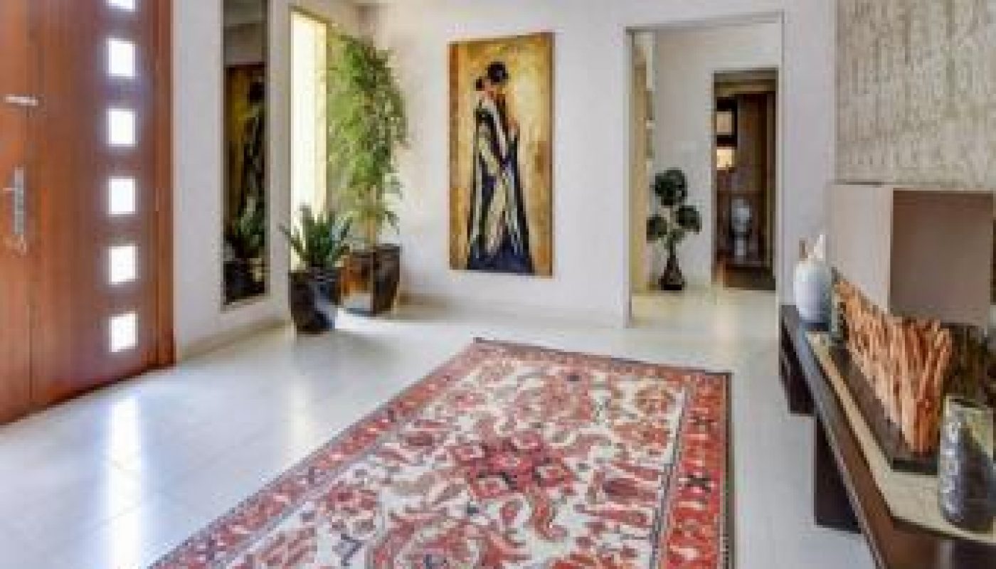Modern entryway with a wooden front door, large rug with red patterns, potted plants, wall art of a woman in a dress, and a console table with decor items.