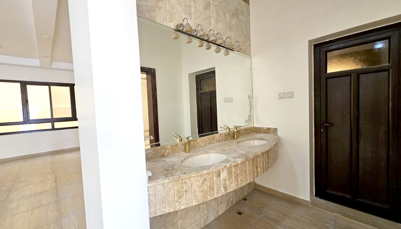 Spacious bathroom with a double sink vanity, large mirror, tiled surfaces, and a dark wooden door. Natural light enters from windows in the adjacent room.
