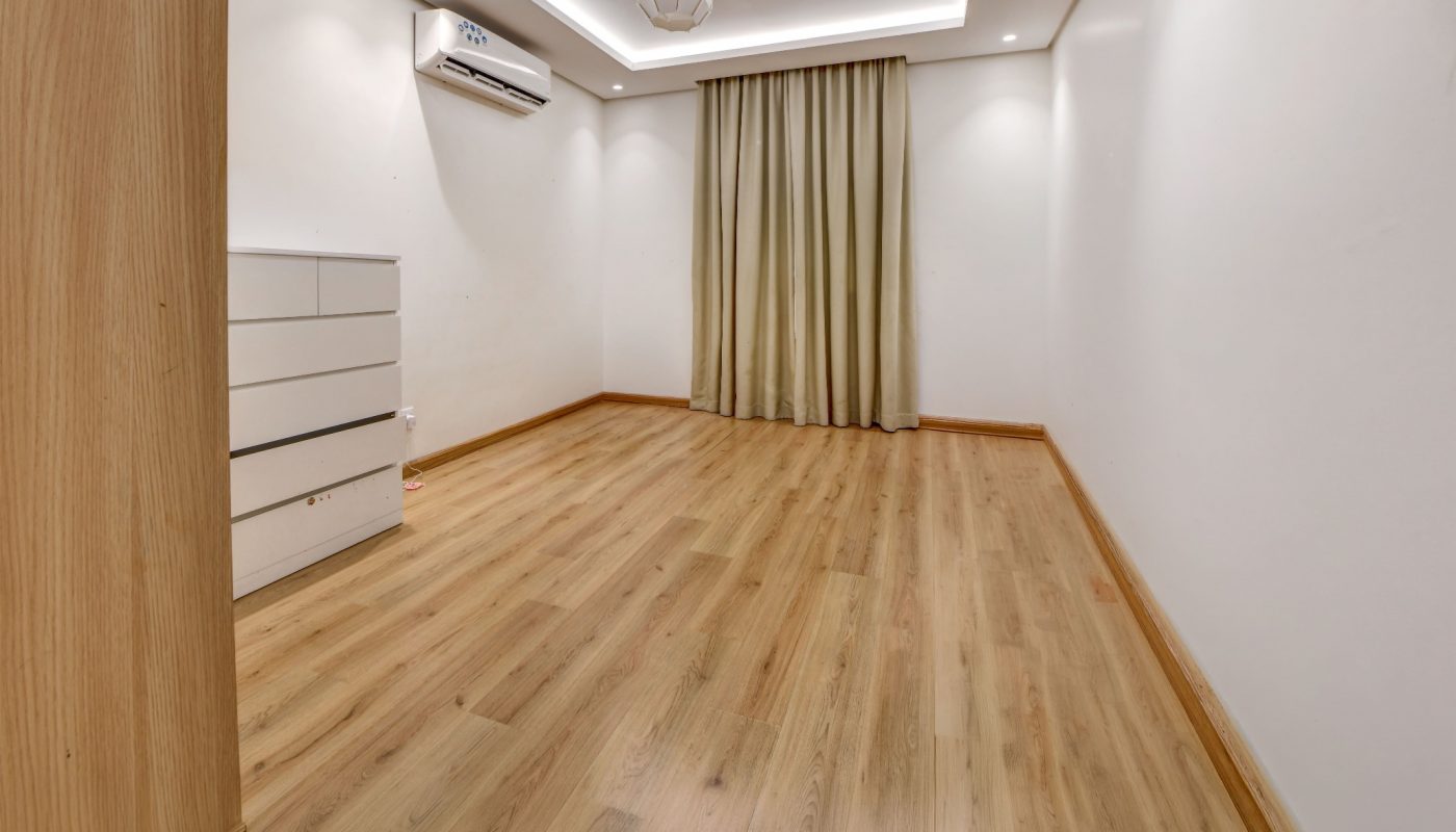 Empty room with wooden floor, white walls, ceiling lights, beige curtains, a white dresser, and an air conditioner mounted on the wall.