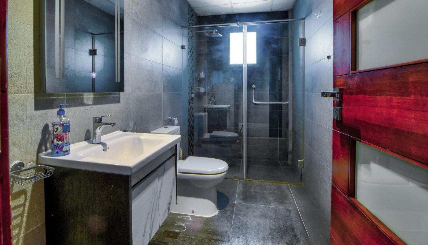 Modern bathroom with gray tiled walls and floors, a wooden door, a large mirror above a double sink, toilet, and glass-enclosed shower with a window.