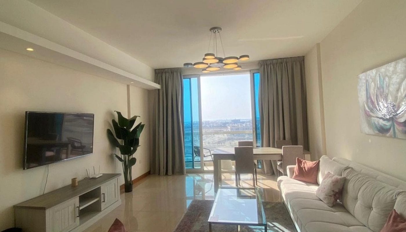 A modern living room with a white sectional sofa, glass coffee table, wall-mounted TV, and large window offering a view of a cityscape.