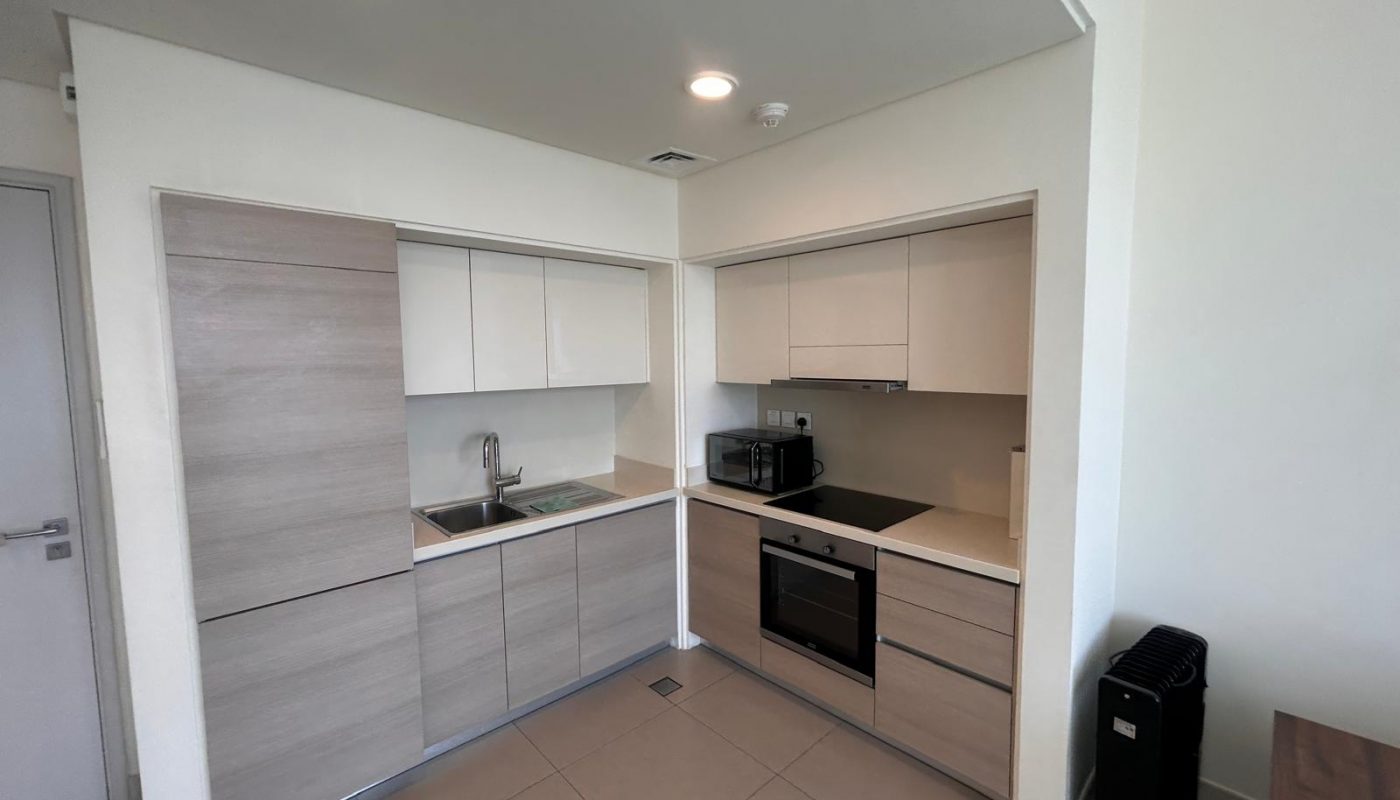 Modern kitchen with light wood and white cabinets, built-in oven, electric cooktop, microwave, and sink. Tiled floor and minimal countertop appliances.