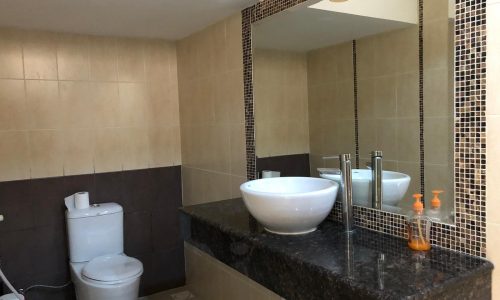 A modern bathroom featuring a large mirror, a white vessel sink on a dark granite countertop, a faucet, a soap dispenser, a toilet, and beige and brown tiled walls.