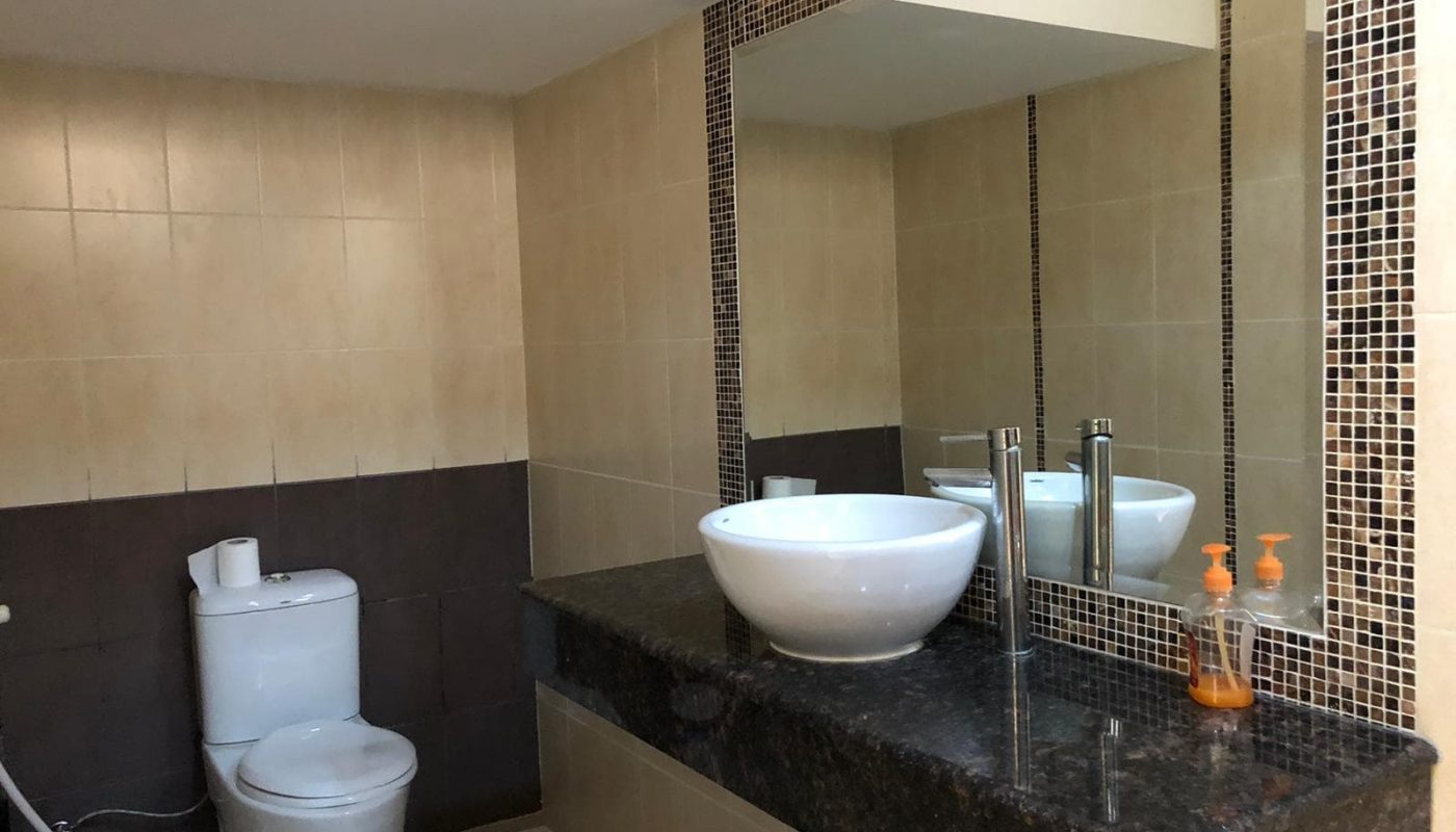 A modern bathroom featuring a large mirror, a white vessel sink on a dark granite countertop, a faucet, a soap dispenser, a toilet, and beige and brown tiled walls.