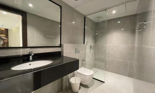 Modern bathroom in an AMWAJ flat for sale featuring a large mirror, black countertop with a sink, toilet, white wastebasket, and a glass-enclosed shower.