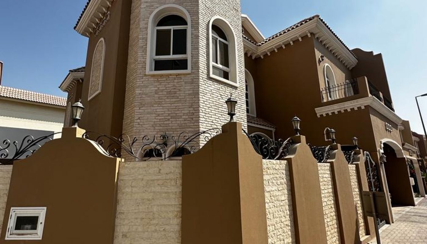 Two-story house with a tall turret and arched windows, surrounded by a brown and beige fence along a paved sidewalk under a clear blue sky.