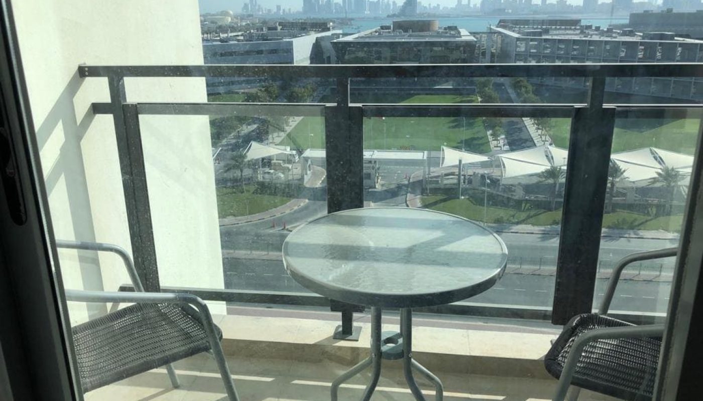 A small balcony with a round glass table and two chairs overlooks a cityscape with buildings, green spaces, and water in the background.