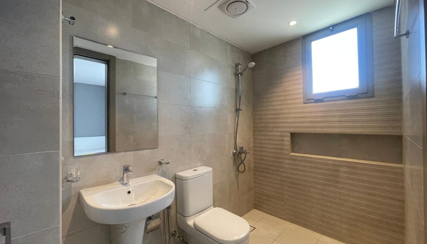 Modern bathroom with gray tiled walls, a wall-mounted sink, toilet, a shower area with a built-in shelf, and a window providing natural light.