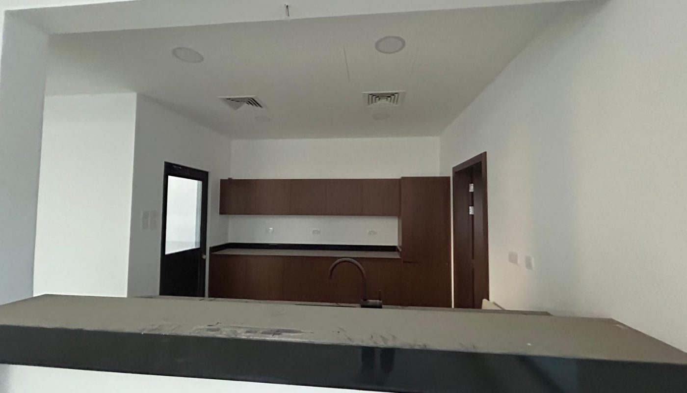 Modern kitchen with dark countertops, wooden cabinets, white walls, and a black faucet, viewed from an open counter window. Two doors and ceiling vents are visible in the background.