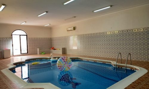 Indoor swimming pool with colorful plastic beach ball floating on the water. Pool area surrounded by patterned tiles and lit with overhead lights. Door and window visible in the background.