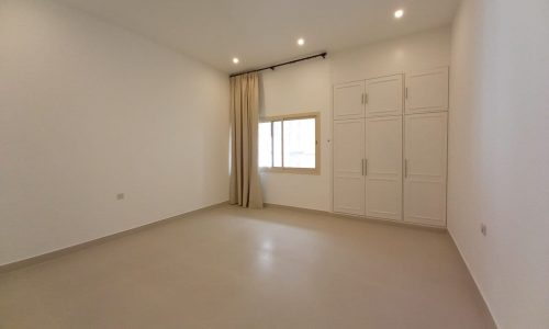 An empty room with white walls, beige tiled floor, a window with beige curtains, and built-in white closets with closed doors.