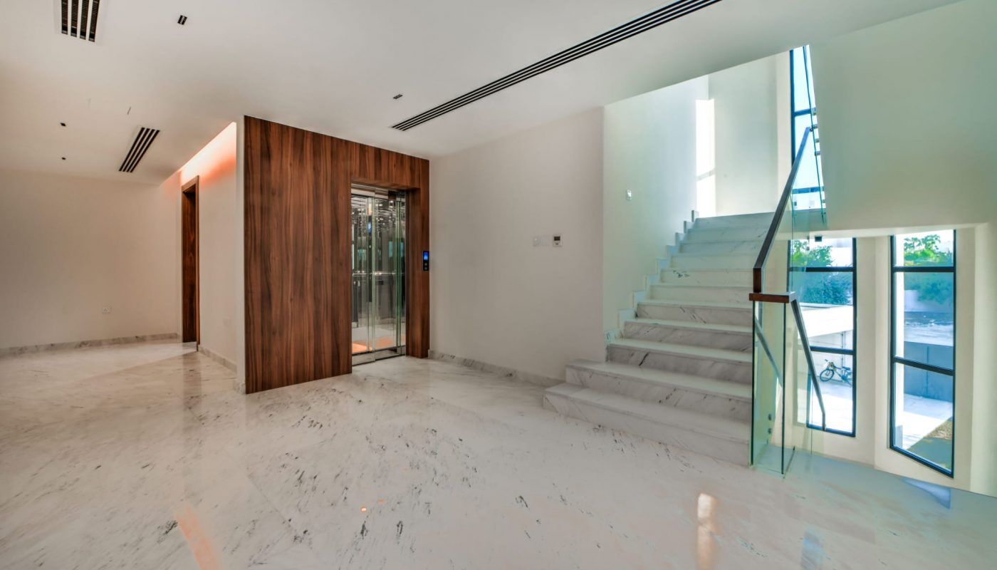 Modern interior with marble floors, a glass elevator with wood paneling, and a white staircase near large windows allowing natural light.