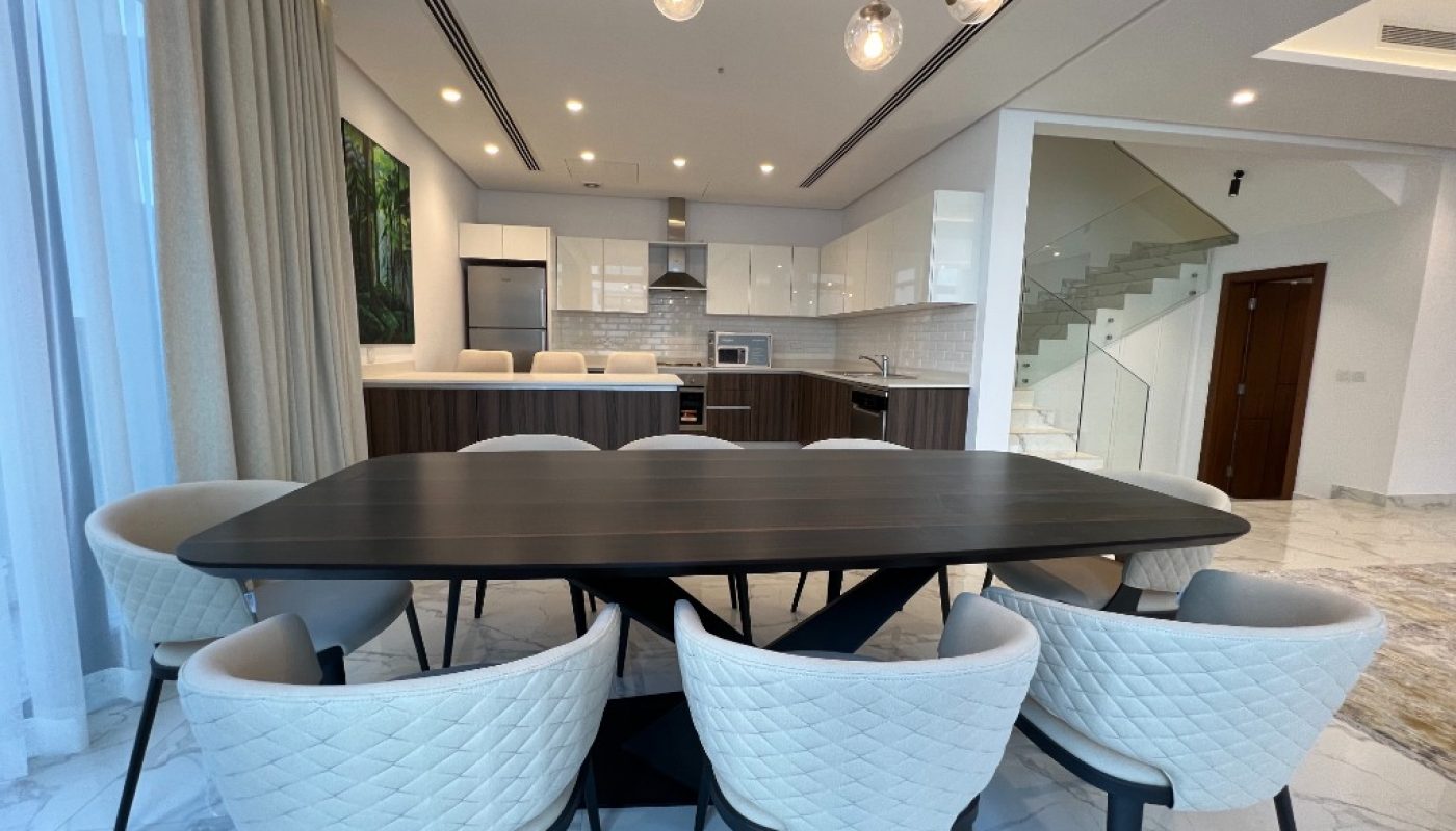 Modern dining area with a dark rectangular table and eight cushioned chairs, adjacent to a kitchen with white cabinets and stainless steel appliances, in an open-plan space.