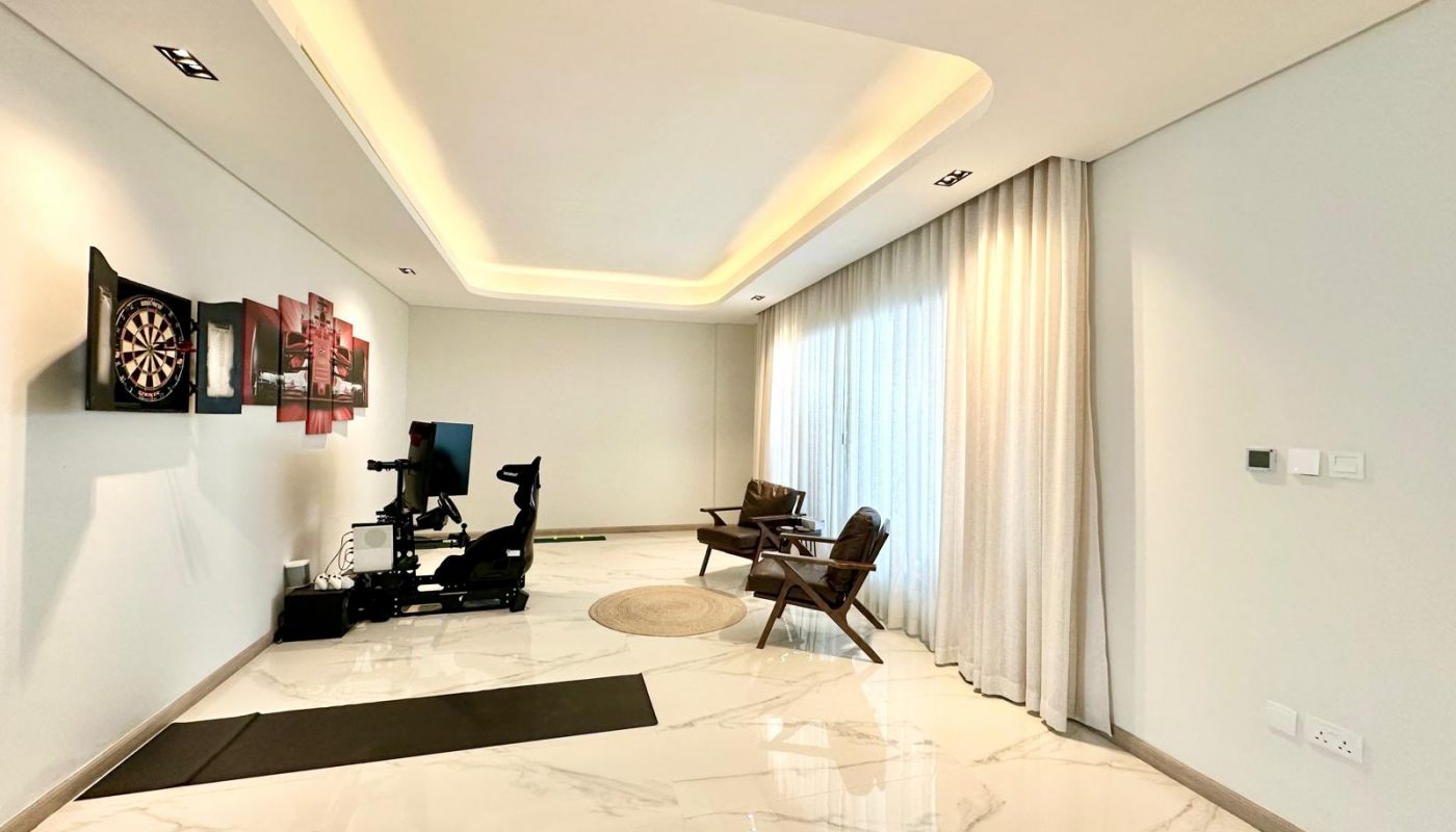 A modern room with marble floors, a dartboard, simulator racing setup, two wooden chairs by large windows with sheer curtains, and recessed ceiling lighting.