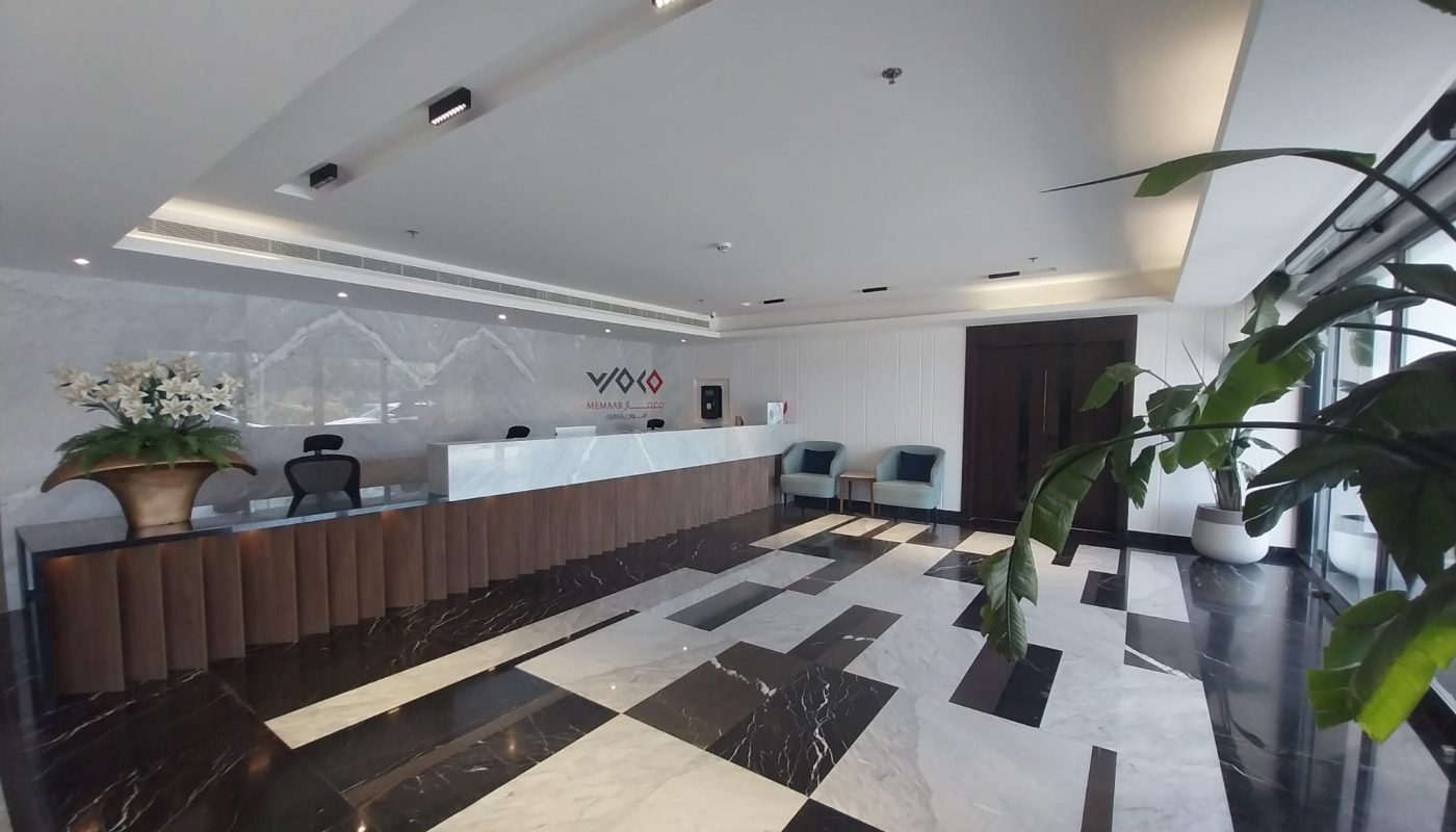 Modern office lobby with a long reception desk, decorative plant, waiting area with chairs, and geometric-patterned flooring.