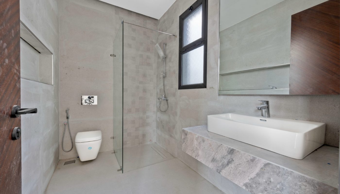 Modern bathroom with a walk-in shower, wall-mounted toilet, rectangular sink, large mirror, and frosted window. The room features minimalist, light gray decor and clean lines.