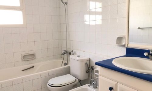 A clean, white-tiled bathroom with a bathtub-shower combo, toilet, bidet, and a sink with a blue countertop and a mirror above it.