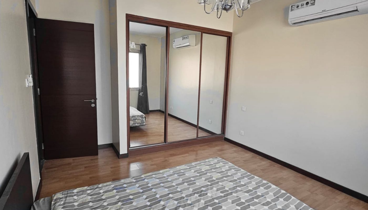 A bedroom with a double bed, patterned bedspread, wooden floor, mirrored wardrobe, dark door, chandelier, air conditioner, and a window with curtains.