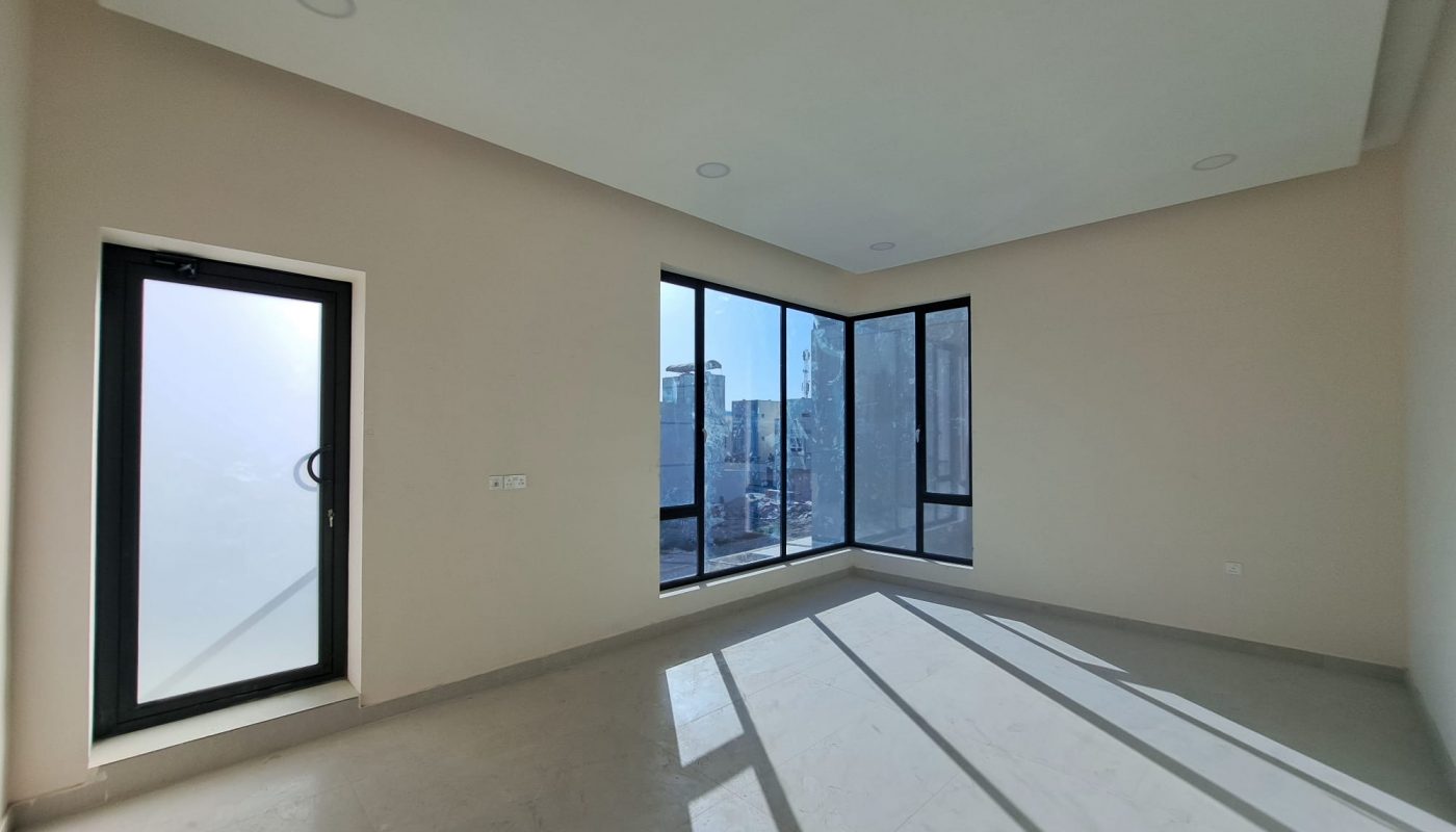 Empty, unfurnished room with beige walls, large corner windows, a single side door, and ample sunlight casting shadows on the floor.