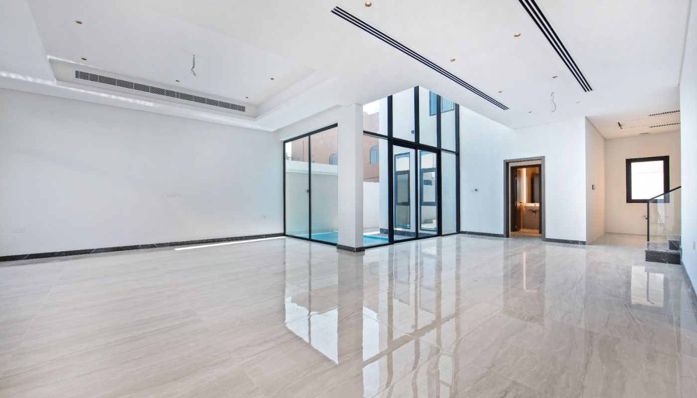 Spacious, empty modern living room with large windows, white walls, marble flooring, and recessed ceiling lights. A glass door leads to a patio; a stairway is visible in the background.