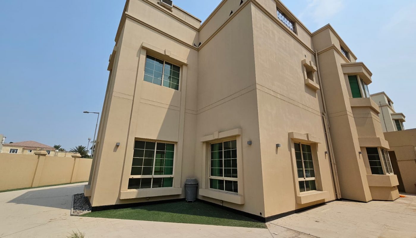 A beige two-story modern house with large windows, flat roof, and a small green lawn, photographed on a sunny day.