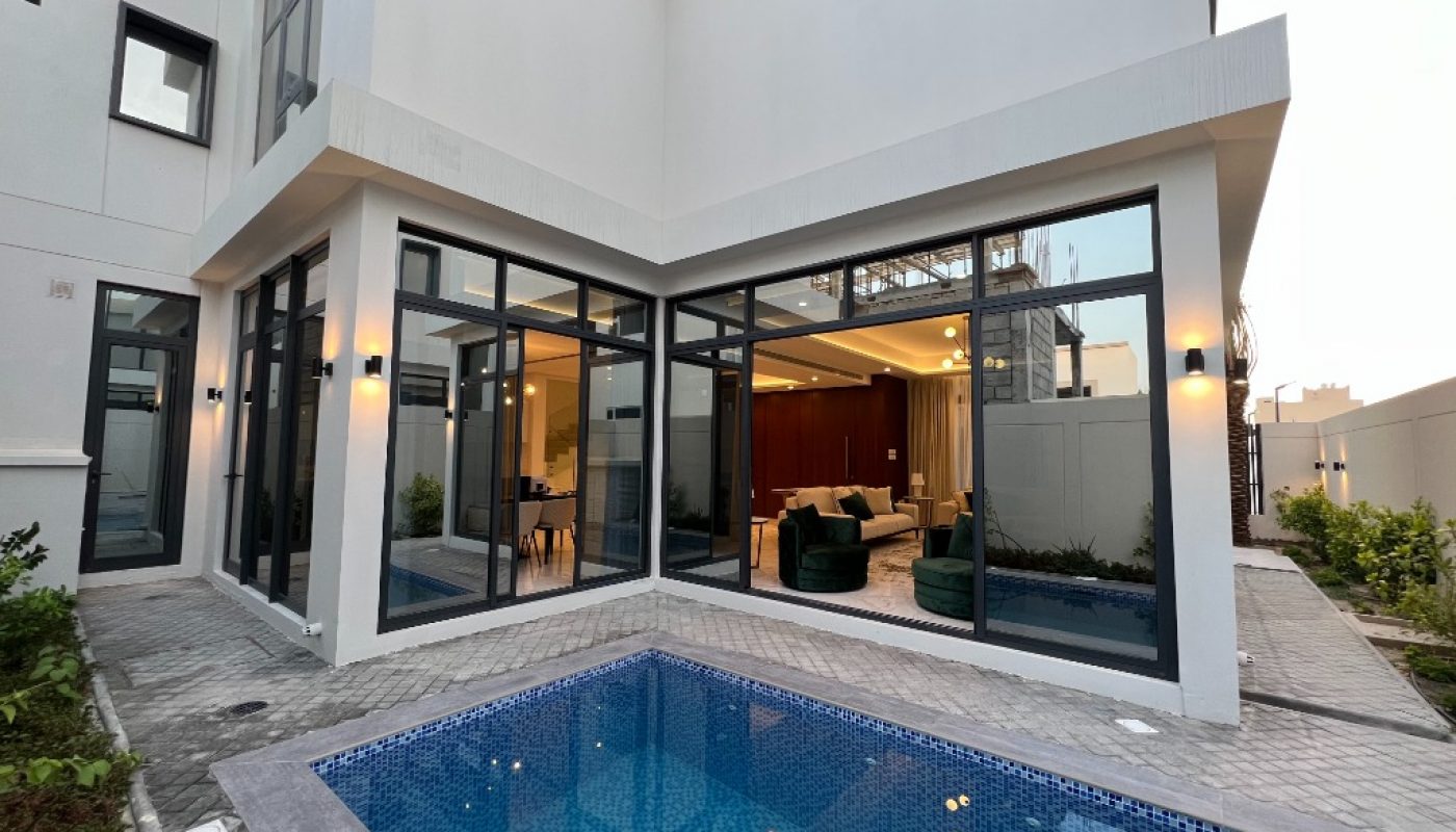 Modern house with large glass windows opens onto a small blue-tiled swimming pool and paved patio, with interior seating visible through the windows.
