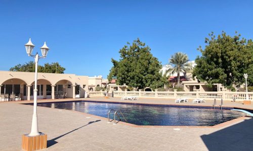 Outdoor swimming pool with clear water, surrounded by a tiled deck and lamp posts. There are several lounge chairs and green trees in the background under a clear blue sky.