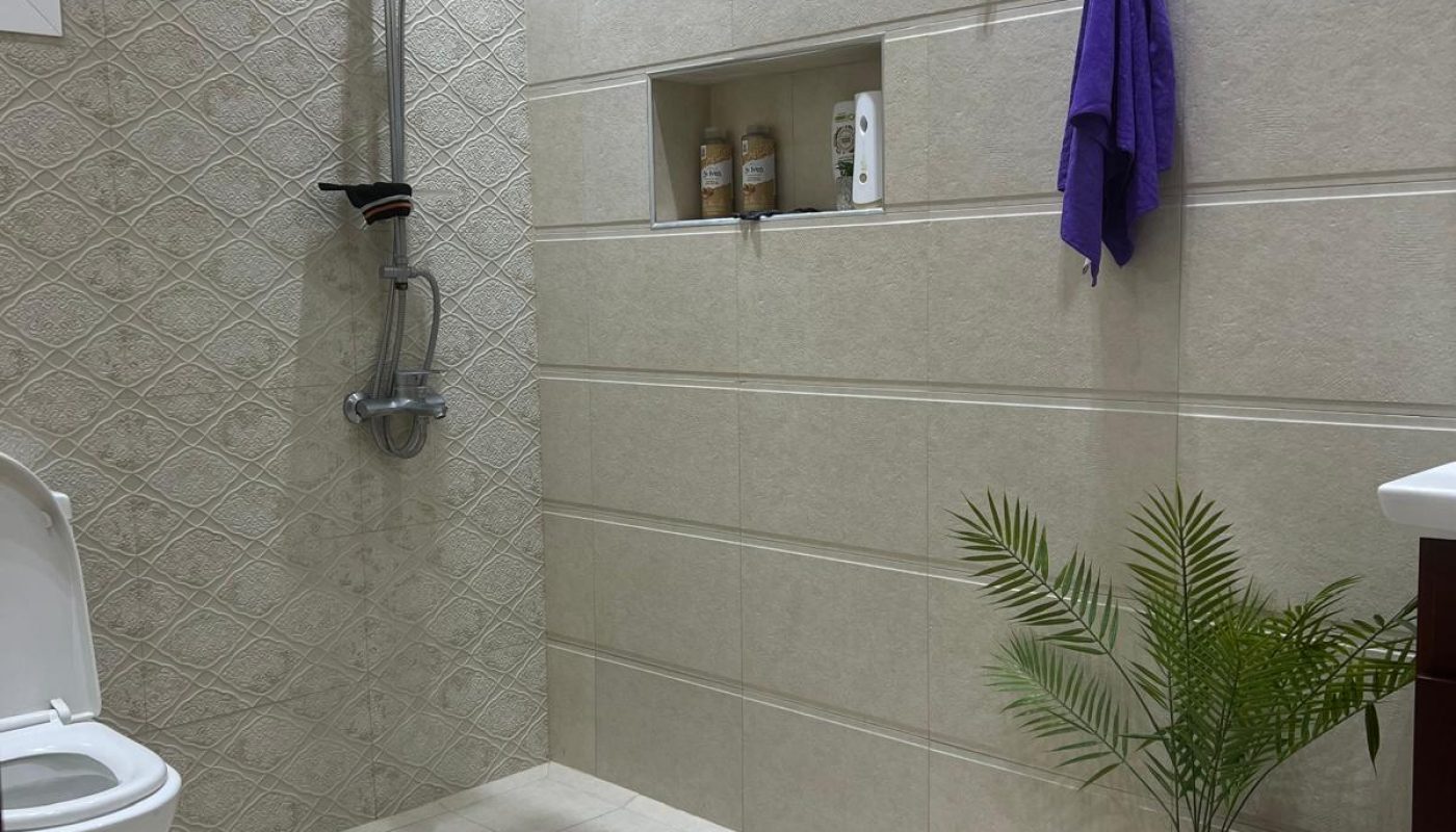 A bathroom with a shower area, a white toilet, a purple towel hanging on the wall, toiletries on a shelf, and a potted plant on the floor.