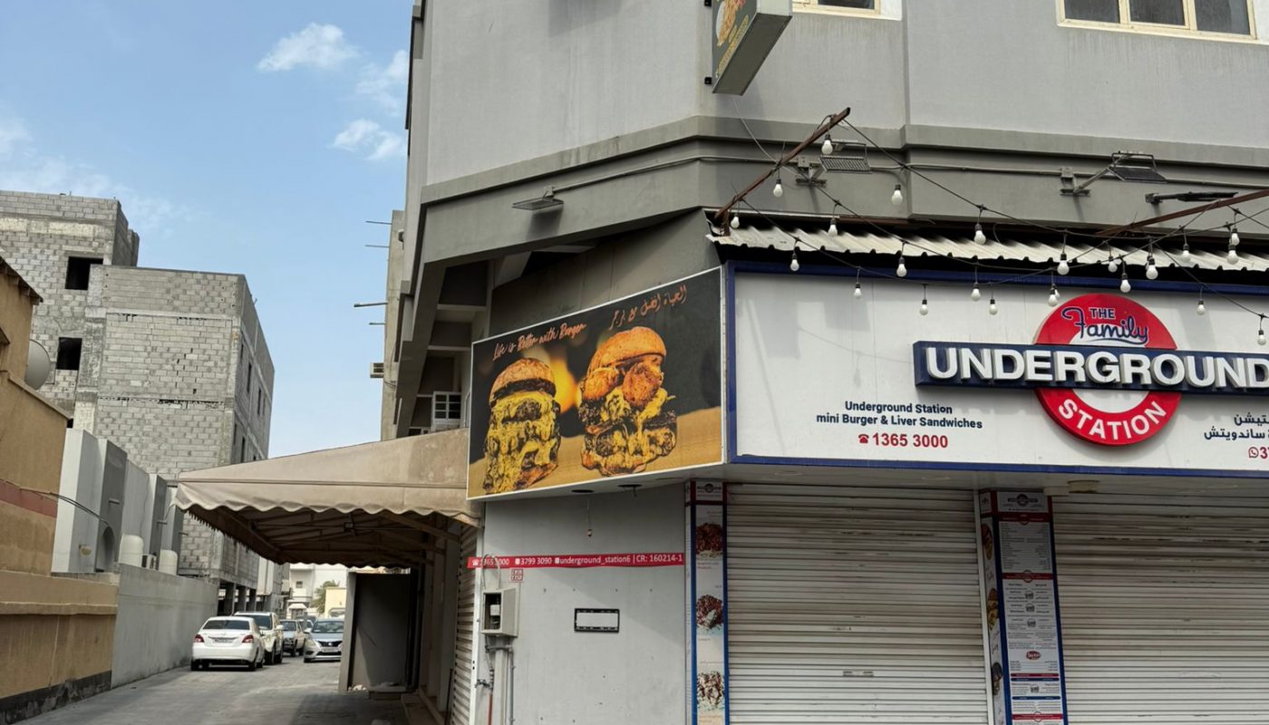 A gray corner building with closed shutters houses a shop called "Underground Station." Above the shop, there is a yellow sign with illustrated faces. The street is empty.