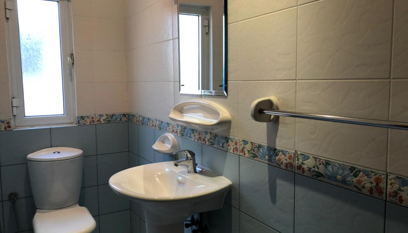 A clean bathroom with a toilet, sink, and mirror in a semi-furnished villa, featuring white and blue tiles and a window on the left.
