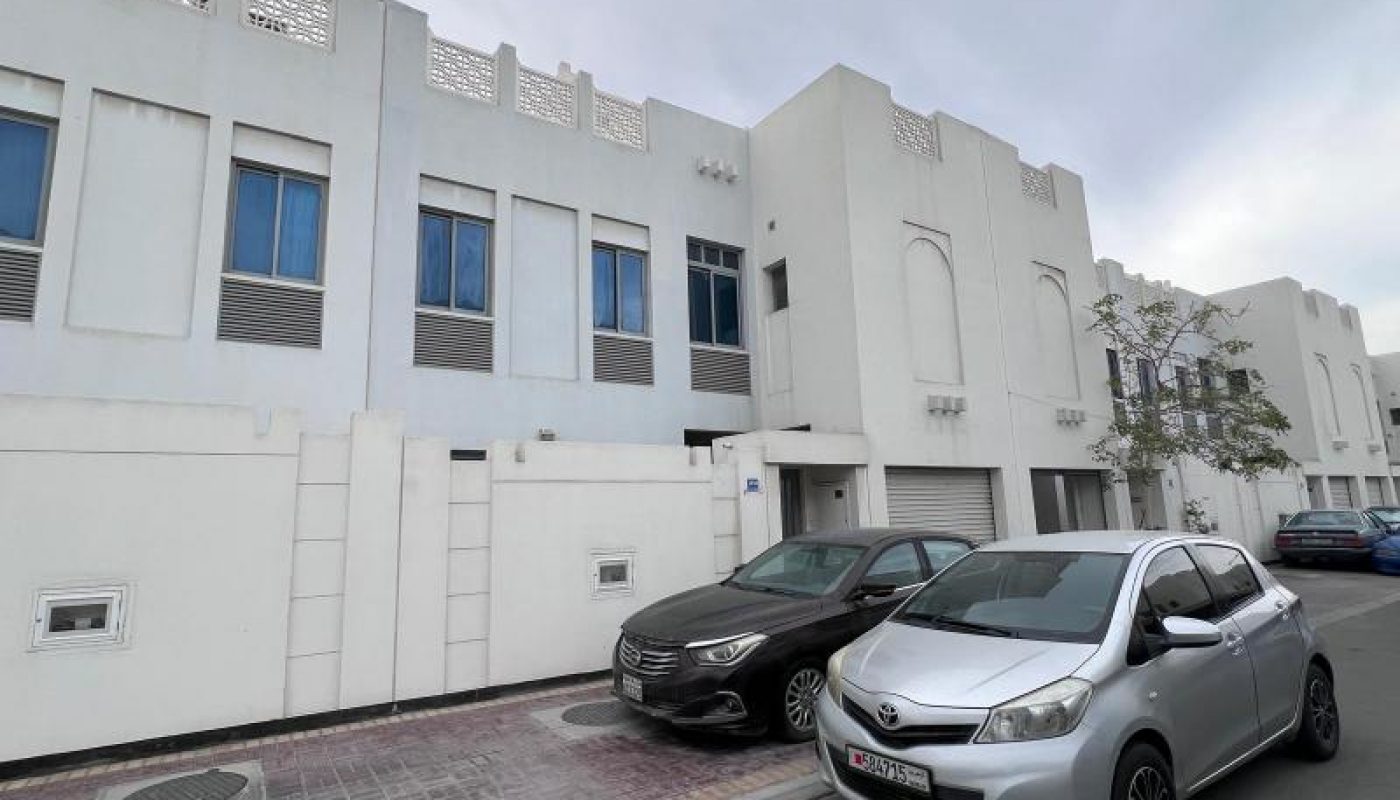 Two cars are parked in front of a white, modern apartment building with blue-tinted windows on a cloudy day.