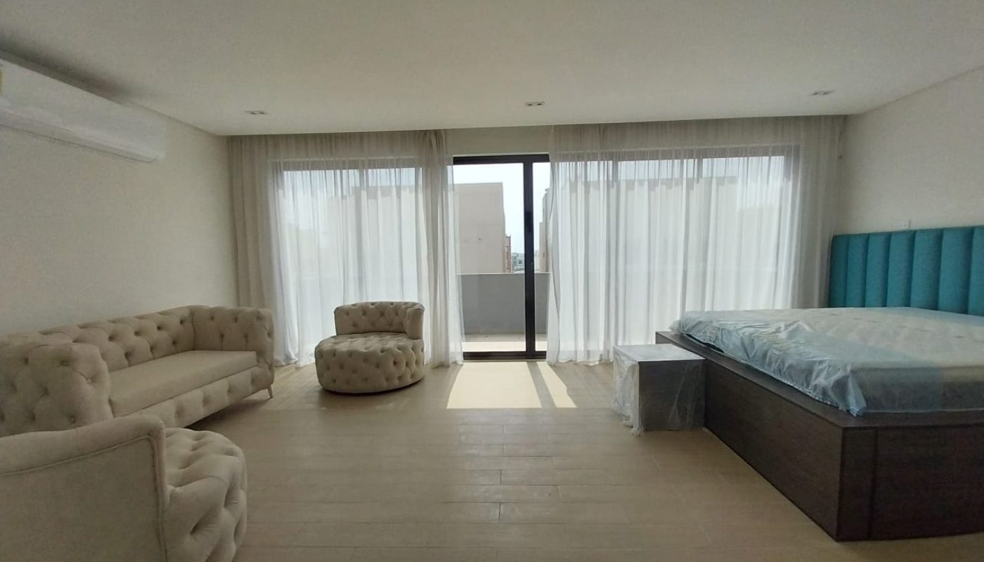 A spacious bedroom with a large bed, beige sofa, and round chair. Floor-to-ceiling windows with sheer curtains provide natural light, and an air conditioner is mounted on the wall.