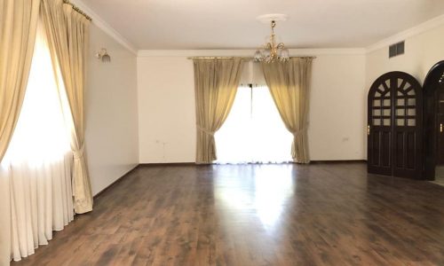 Spacious semi-furnished villa in Saar with an empty living room, wooden flooring, white drapes on windows, and a chandelier hanging from the ceiling.