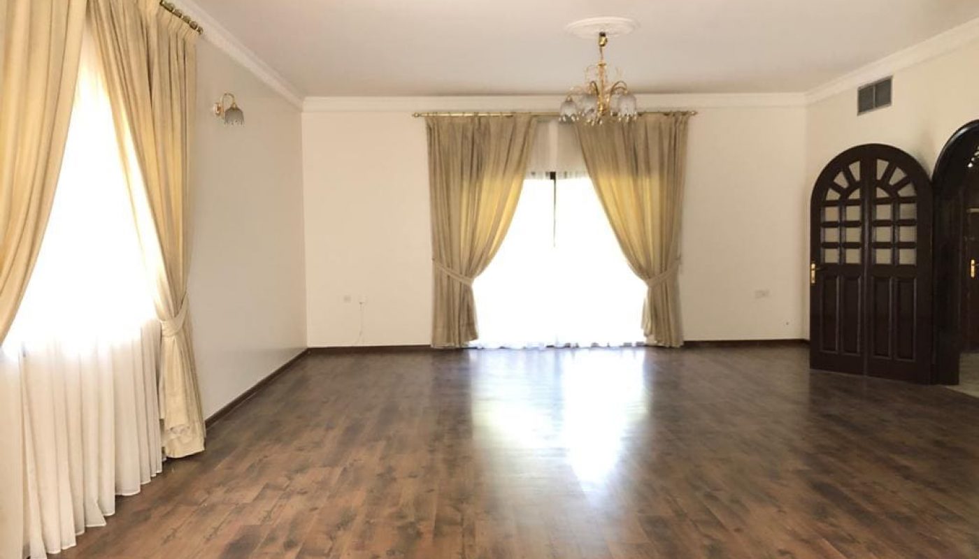 Spacious semi-furnished villa in Saar with an empty living room, wooden flooring, white drapes on windows, and a chandelier hanging from the ceiling.