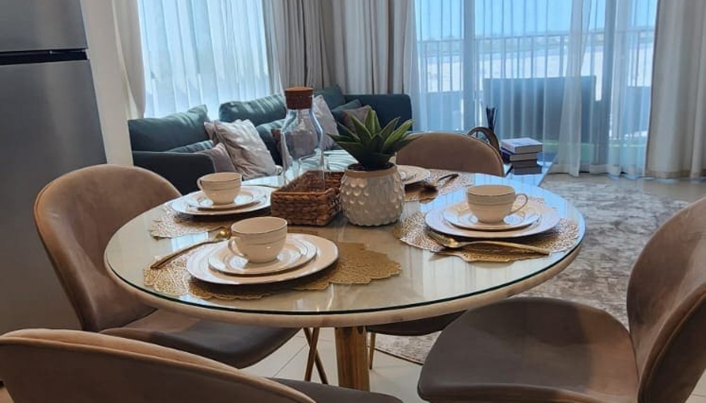 A dining area with a round table set for four, surrounded by chairs. Large windows with sheer curtains offer a view outside. A modern chandelier hangs above, and a sofa is visible in the background.