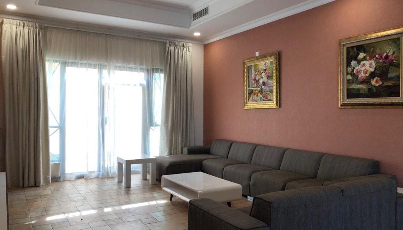 A bright living room in a luxury villa with a grey sofa, white coffee table, tiled floor, pink walls, floral paintings, and curtains partially covering a glass door.