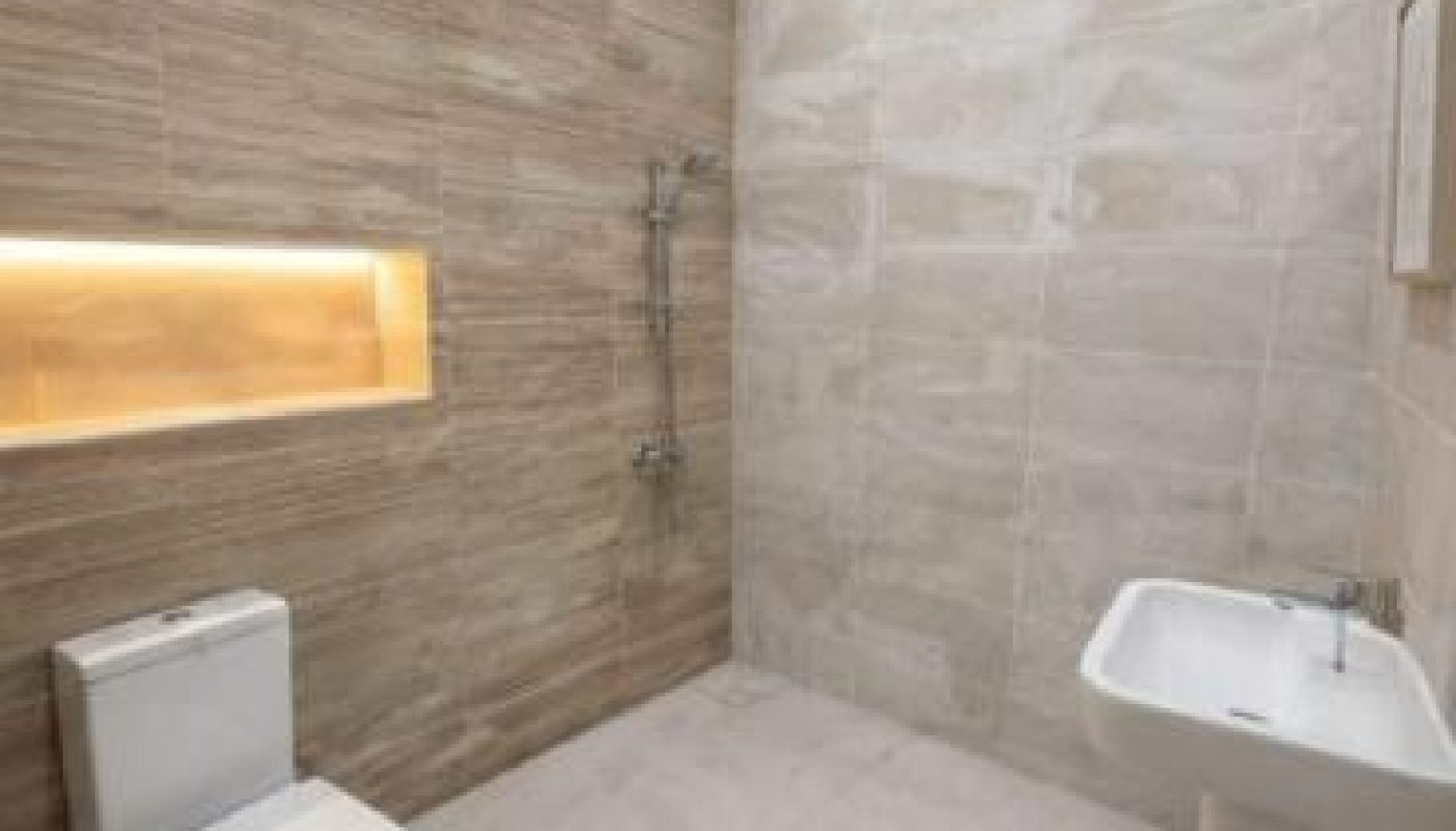 Modern bathroom with a wall-mounted sink, toilet, built-in shelf with lighting, and shower area, featuring beige and light brown tiles.