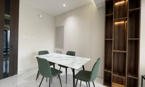 A modern dining room in an AMWAJ flat for sale features a white marble table, four green chairs, and an empty wooden shelving unit under warm lighting.