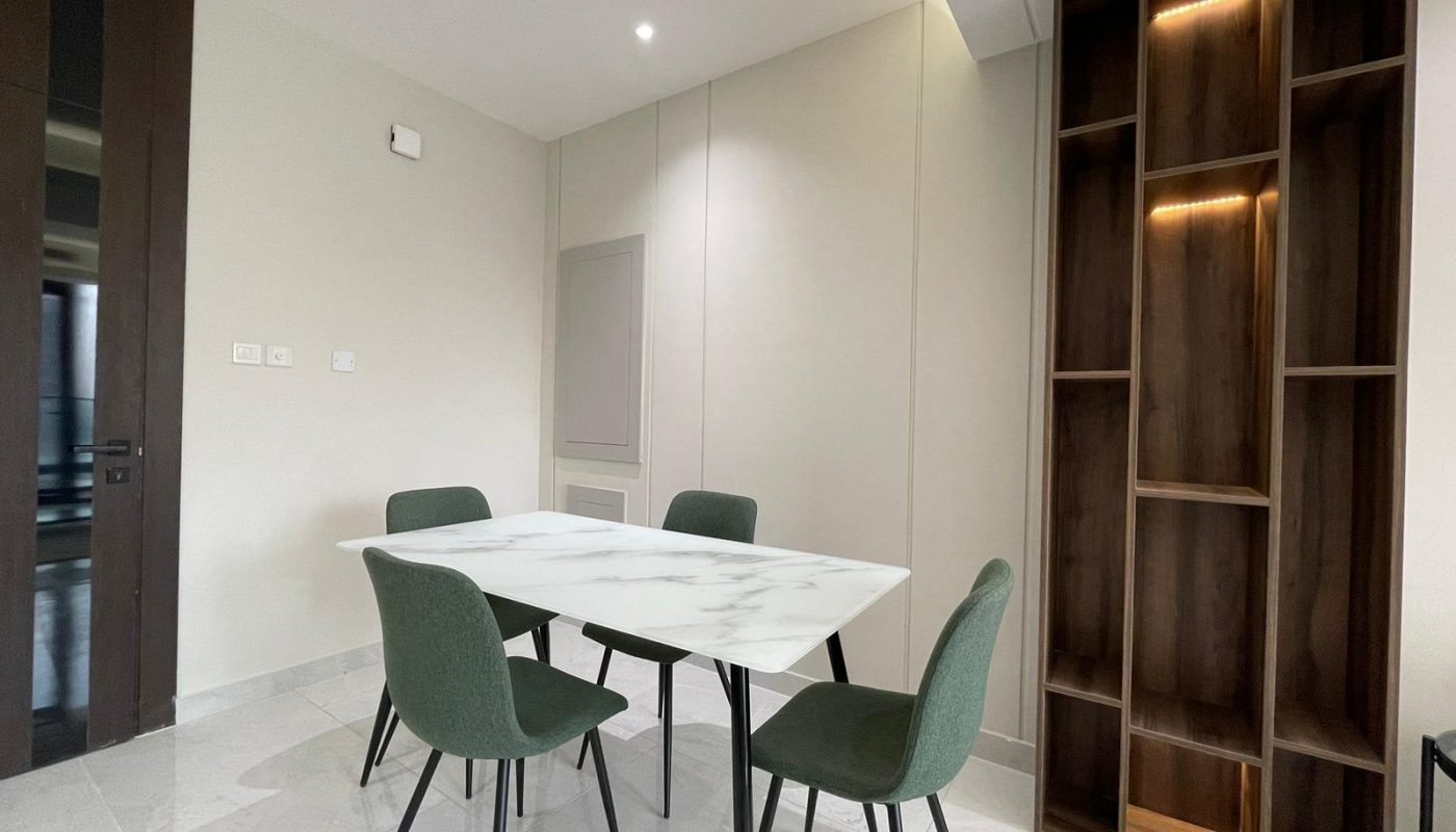 A modern dining area with a white marble table, four green chairs, and an empty wooden shelf with built-in lighting against a light-colored wall.