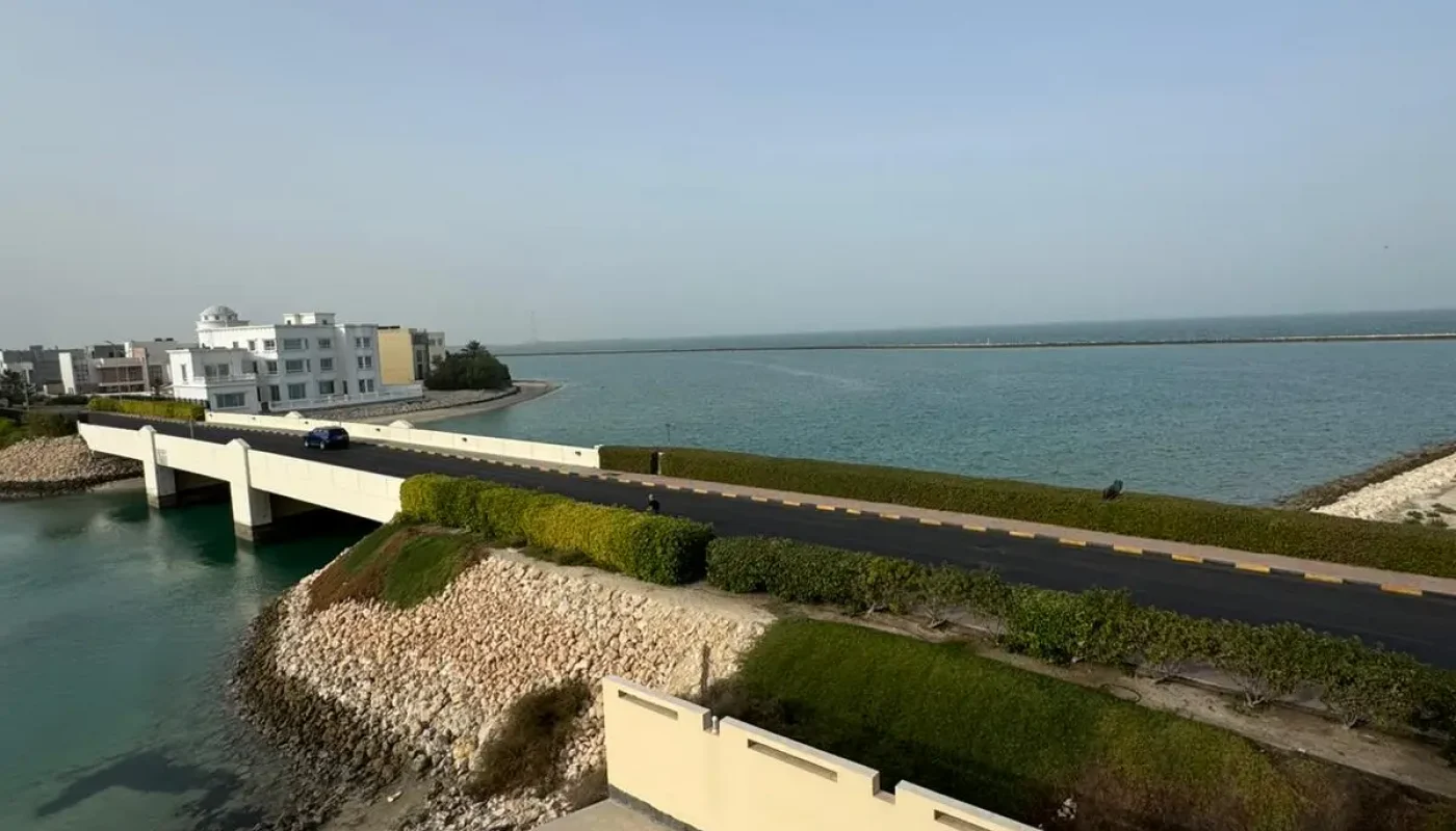 A view of a bridge over turquoise water, with a road and modern buildings on the left, and the sea and sky in the background.