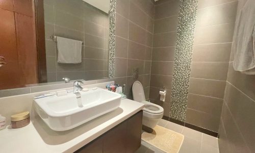 A modern bathroom with grey tiles, featuring a sink, mirror, toilet, and Auto Draft towel rack.