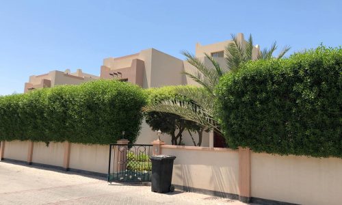 A beige building with a flat roof is partially obscured by a tall, well-manicured hedge and palm tree. A black trash bin stands next to a gated entrance, and a clear sky is visible in the background.