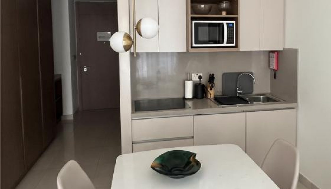 Small modern kitchen with beige cabinets, built-in sink, microwave, and a white dining table set for two with light brown chairs. A decorative bowl sits on the table.