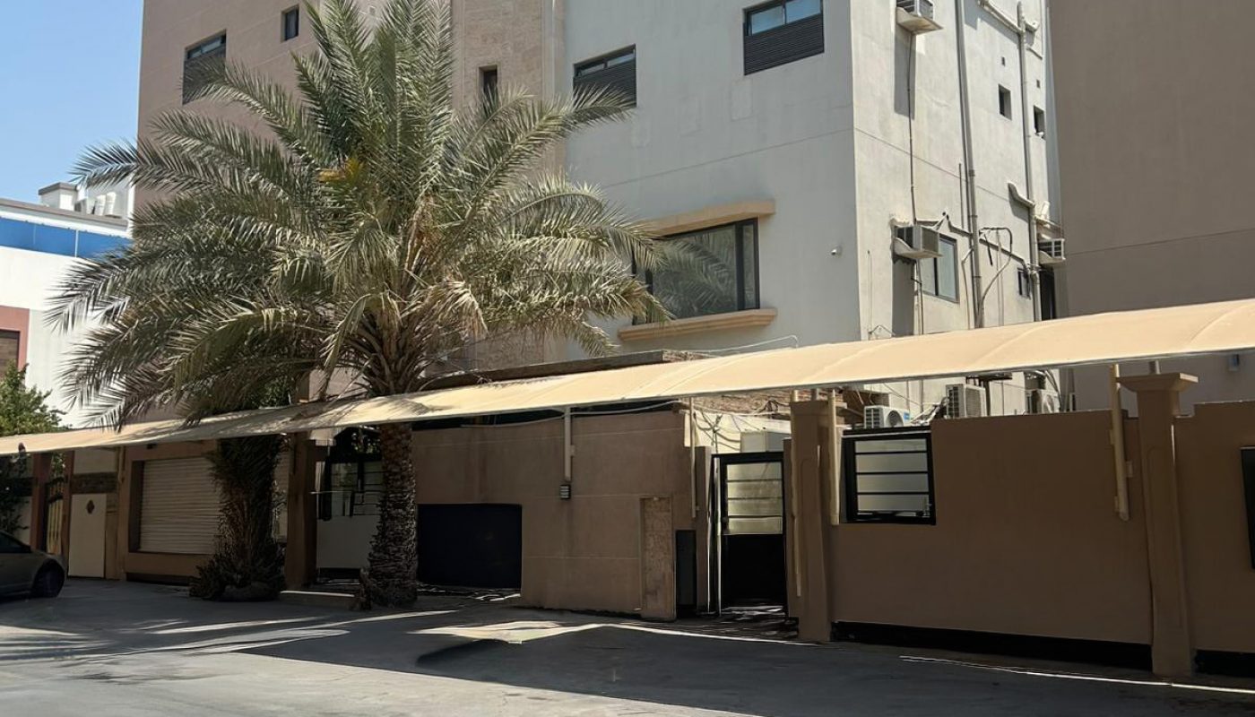 Three-story beige residential building with covered parking spaces, palm trees, and a clear blue sky in the background.