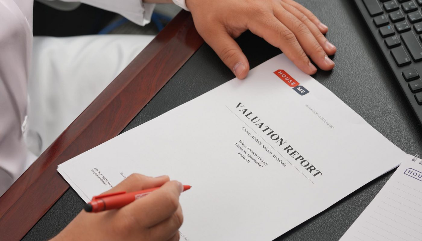 Person holding a red pen sits at a desk with a valuation report document, wearing a smartwatch, next to a keyboard and a notepad.